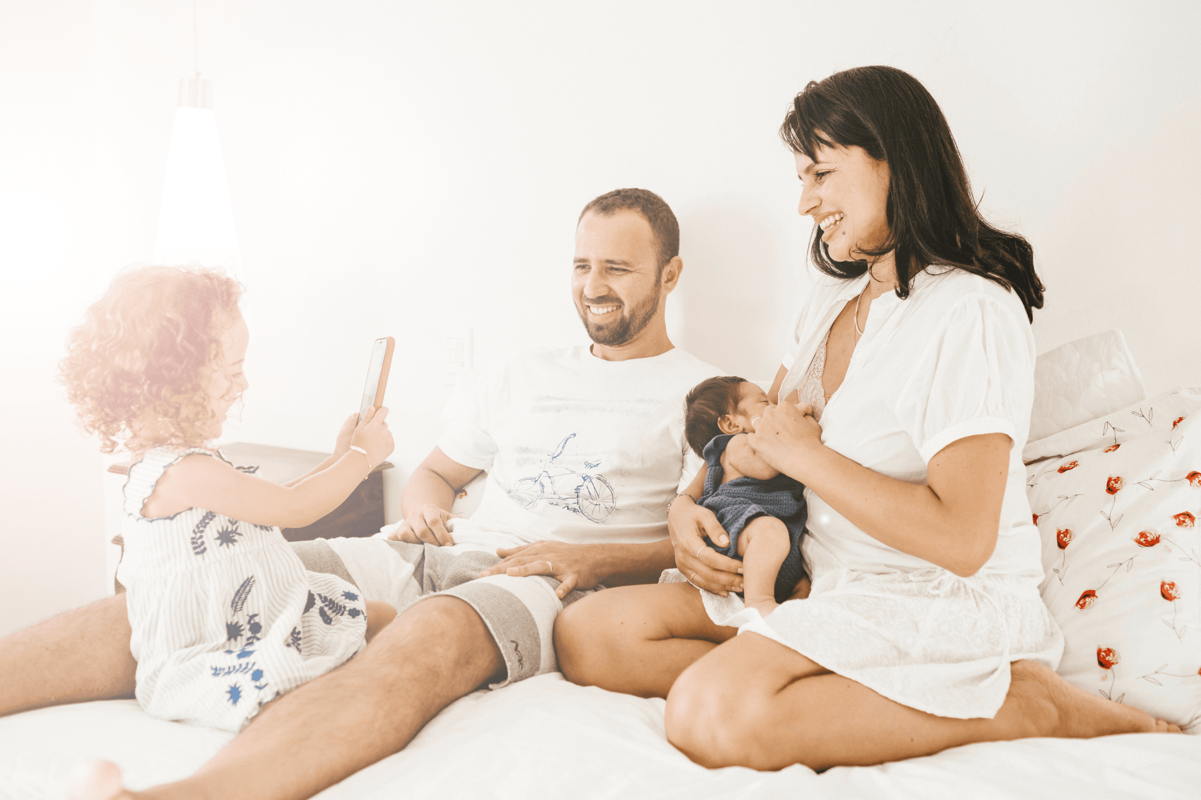 A happy family sitting together on a couch, with a mother holding a baby and a father smiling at a child taking a photo with a smartphone.